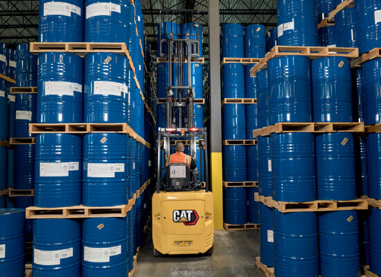 Worker driving Cat cushion lift truck through narrow aisle of stacked barrels